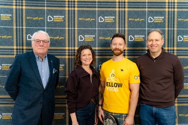 Four people smiling in front of a tartan Beatson Cancer Charity backdrop, one wearing a yellow Yellow Yogis T-shirt.
