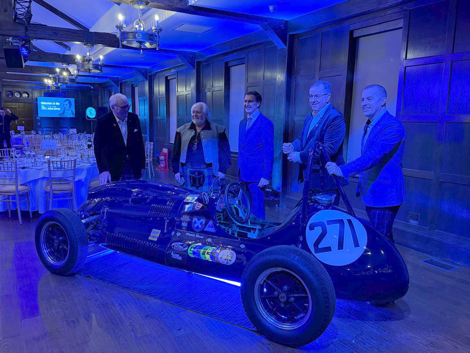 Barry Wood, far left, and four other men in suits and tartan standing behind a dark blue 1950s single-seater racing car, number 271, in a wood-panelled dining room lit in blue and set for dinner.
