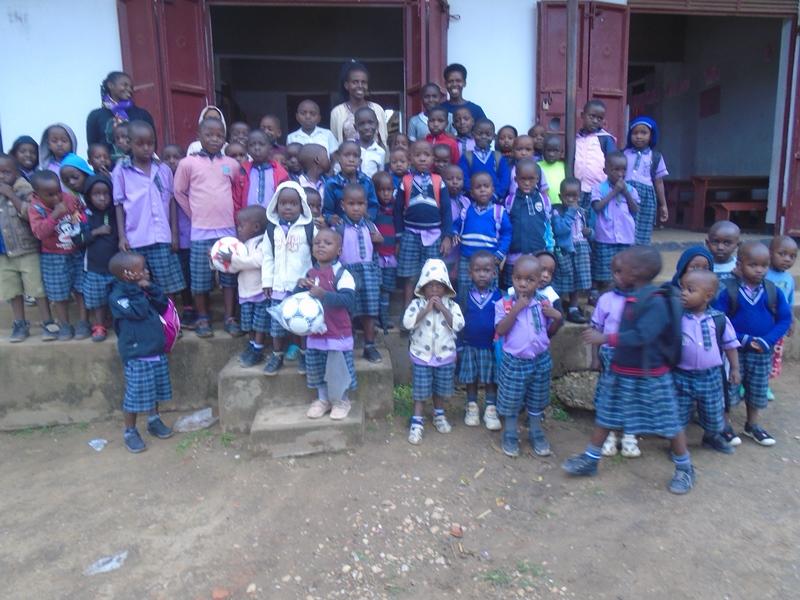 A large group of children in purple and navy school uniforms standing outside Ruth Memorial Orphanage School in Uganda, some holding footballs.
