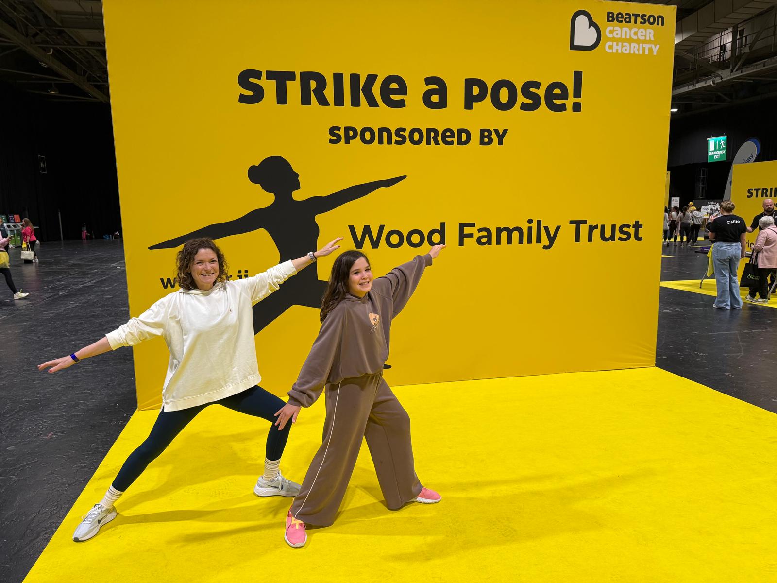 Niamh and her daughter doing a yoga warrior pose on a bright yellow stage, in front of a Beatson Cancer Charity sign reading ‘Strike a pose! Sponsored by Wood Family Trust’.